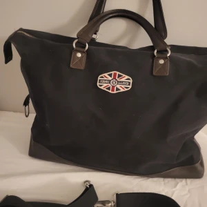 Henri lloyd weekendbag - En stilren och snygg weekendväska ifrån henri lloyd. Små slitage förekommer. Trådarna på fästet i ena sidan av väskan håller på att lossna. Se bild 3. Jag har tyvärr ingen nål och tråd så jag kan fixa det.  Originalkedja är bytt till ersättnkngskedja. 35L