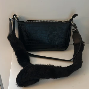 Svart väska  - En svart vardaglig väska. Köpt för dryg ett år sedan. Väskan är i bra skick då den använts fåtal gånger. Det fluffiga bandet har jag köpt till själv, men man får med den också! 