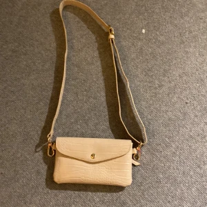 Handväska  - En begie handväska med gulddetaljer och justerbar axelrem. Rymmer en mobil och ett eller två kort. En pytteliten fläck på insidan dom antagligen går bort om väskan tvättas. Den är köpt på Lindex. 