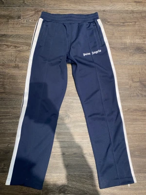 Palm angels taped track pant - Palm angels taped trackpants.  Säljes för att dem är lite för stora för mig.  Storlek: Large Skick: 9,5/10 Navy blue  Köpta nya i september för 4295kr och inte använda mycket alls.  Inga flaws. Box påse tags och digitalt kvitto finns. 