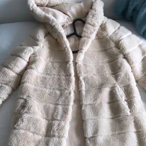 Fuskpälskappa - Fuskpälskappa med huva. Två öppna framfickor och Längd: 78 cm i storlek S Bra i skick och inga repor.