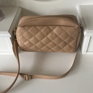 Liten handväska - Liten beige handväska med justerbart axelband och även en ”guld” detalj på framsidan. Väskan är aldrig använd och blixtlåset funkar perfekt. Väskan är köpt från Shein. Kan skickas omgående, pris kan diskuteras.