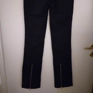 Svarta smala byxor med dragkedjor på benen, från ATT/ÅHLÉNS. Strlk 34. - Svarta smala byxor med silverfärgade dragkedjor på benen, från ATT/ÅHLÉNS. Strlk 34. Begagnade men inte trasiga etc. Stängs med dragkedja och knapp. Material: Bomull.