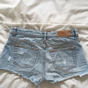 Lågmidjade jeansshorts - Ljusblå lågmidjade jeansshorts i Storlek XS. Midjemått 36cm, innerbenslängd 4cm. 100%  bomull DEFEKT: slits i höger ben, se bild 3. Slitningen är cirka 4cm lång och utgör ingen större skillnad för hur shortsen ser ut då de redan har en sliten look.