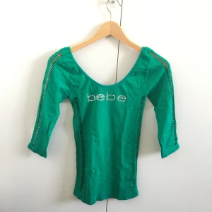 Topp från Bebe - Trendig grön tröja från märket Bebe, stark 00-tals stil! Med text i stenar över bröstet och ett mönster av hål längs sidan och ärmarna. Stretchig och tight så passar fler storlekar! Mått: 29.5 cm mellan armhålorna. Längd 59.5 cm från axeln och ner.