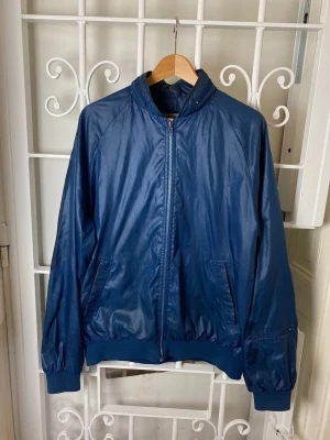 Vintage-jacka från Duckster - Plastig blå fritidsjacka från klassiska klädföretaget Duckster.  Vintage i mycket fint skick.  Huva i blixtlås vid nacken, innerficka och två fickor med blixtlås. 100 % nylon.