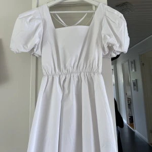 Vit klänning  - Vit knälång klänning med snörning i ryggen och puffärm. Resår i midjan. Lite tjockare tyg. Perfekt för student eller skolavslutning. Helt ny. Endast provad. Lappen sitter kvar. 
