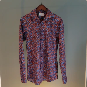 Skjorta  - Blå/orange skjorta med blommigt mönster. Passar till att ha under en pullover samt vara skjortan i sig själv. Passar till de flesta av tillfällen. 
