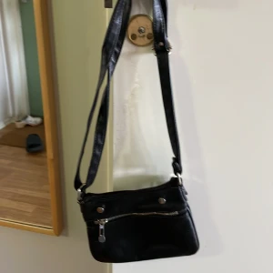 Axelremsväska  - Man kan spänna åt den. Det är en dragkedja.