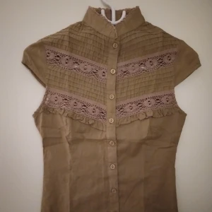 Vintage look, brun kortärmad broderad blus. Strlk: XS, Petite. Köpt utomlands.OANVÄND. - Vintage look, brun kortärmad broderad blus med knytband/snörning i ryggen. Strlk: XXS-XS, Petite. Köpt utomlands. OANVÄND. Helt felfri. MÄRKET ÄR EJ CANADA GOOSE, DET ÄR TEKNISKT FEL AV PLICK SOM EJ GÅR ATT ÄNDRA, MÄRKLIGT NOG. 