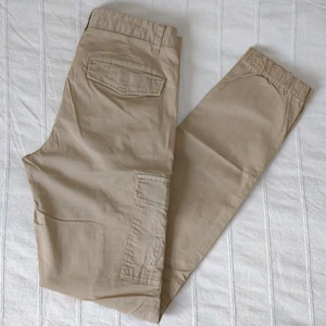 Beigea cargobyxor  - Beigea cargobyxor med stretch. Lite resår i bensluten. Fickor på benen och i bak - med knappstängning under locket. Använda bara ett par gånger och i fint skick. 98% bomull och 2% elastan. Storlek 36.