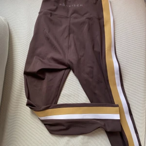 Röhnisch träningstights med hög midja  - Röhnisch träningstights med hög midja och snygga detaljer!   Tightsen är i mörkbrun färg med snygga detaljer på sidorna och liten ficka inuti bak.  Tightsen är lite tjockare och i väldigt ”slittåligt” material.  Byxorna är endast använda 1 gång.   