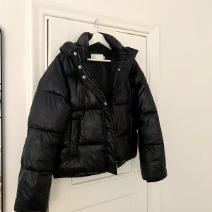 NLY TREND short puffer jacket Strl 34 - Svart, kort vadderad jacka med huva i ett glasaktigt material. Strl 34. Mycket gott skick. Slutsåld på hemsidan. Inköpt för 479 kr. 