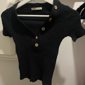 Svart Pull & Bear t-shirt - Svart Pull & Bear t-shirt i ribbat tyg med knappar på, i storlek S men kroppstajt passar även XS. Använd några gånger men ser gott som ny ut.