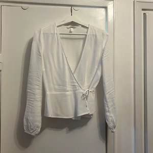 Jacuardvävd omlottblus, H&M stl. 36 - Skir vit blus, med två knytningar, har en hyska upptill i V-ringningen för ytterligare knäppning. Tunn söm som markerar midjan och självfall i tyget under sömmen. 