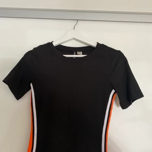 Klänning  - En tajt men stretchig klänning som slutar en bit över knäna i storlek S. Den är svart med ett orange och ett vitt sträck på båda sidorna av klänningen. Den är bara använd ett fåtal gånger och i väldigt fint skick🥰 köparen står för frakten men kan också skicka den via post!