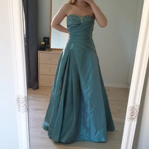 Blågrön balklänning - Blågrön balklänning. Pärlor högst upp på klänningen och samlat i midjan. Jag är 165. På första bilden står jag på tå, därmed kan dem som är ungefär 165 använda klackskor eller sy upp den med en söm eller dem som är längre kan ha plattare skor. Jag är vanligtvis 36. På mig sitter den lite stort i midjan (jag håller in klänningen på första bilden) och mer stort över brösten. Skulle säga att den passar bra till 36/38. Märket är Alicja Eklöw