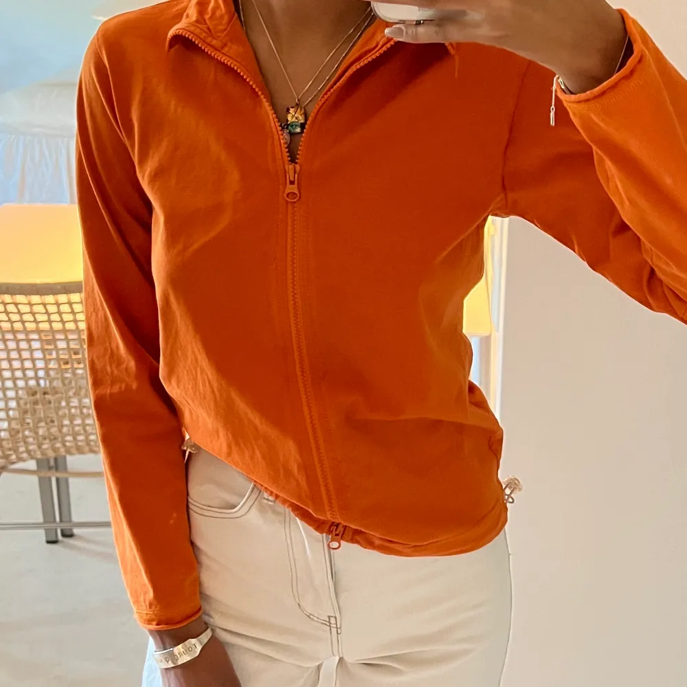 En supersnygg orange tröja köpt second hand från Humana. Tröjan har en dragkedja som kan förslutas både upp- och nedifrån vilket gör att den antingen kan stängas helt eller lämnas öppen. Plagget kan även tajtas till nedtill. Passar de med storlek XS/S. Tröjor & Koftor.