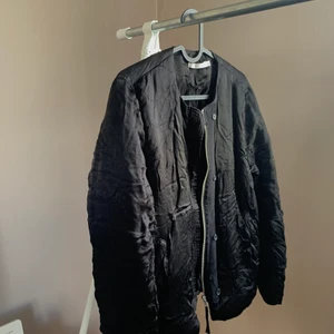 Jacka från WERA i strl 38 - Svart ish bomberjacka i skimrande tyg från WERA i strl 38. Jättemjuk och skönt material. En lös tråd för fäste av knapp som den händige säkerligen kan fixa, dragkedja finns. Såg vid granskning en tråddragning på baksidan samt en liten vi fläck vid axel
