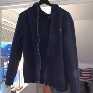 Marinblå Ralph Lauren zip hoodie som är sparsamt använd, den är i nyskick! Det är juniorstorlek XL, vilket motsvarar storlek S i herrstorlek. Nypris: 1100kr