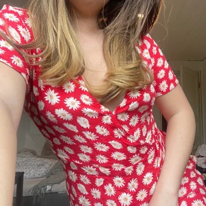 Röd sommarklänning med blommor - Supersöt kort röd  omlottklänning med vita blommor och knyte i midjan. Jätteskönt material och i nyskick. Hör av dig vid intresse eller frågor!❤️