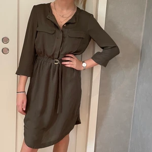 Grön klänning - En fin klänning som passar året runt. Sparsamt använd, är som ny. Fina detaljer med enkelt bälte, knappar och ficklock. Normal i storleken. Trekvarts ärmar. 