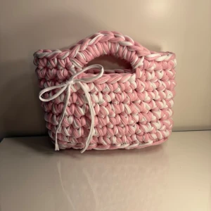 Virkad handväska i t-hirt garn i rosa/vit - •Virkad handväska som smyckas av en söt rosett  •Virkad i t-shirt garn •Färg: Rosa & Vit   •Storlek S Mått: ca 20 cm bred & ca 18 cm hög  •Obs: Storlekarna kan skilja lite,  beroende på garnets tjocklek.  •Virkad av mig: @virkningbynadja   •Kolla gärna in min Instagram, @virkningbynadja 