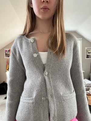 Grå ribbstickad kofta med fickor - Säljer en stilren grå ribbstickad kofta med rund halsringning och stora silvriga knappar. Koftan har två praktiska fickor framtill och är långärmad. Perfekt för en stilren men avslappnad outfit