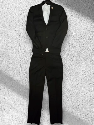 Svart kostym dressmann med kavaj skjorta och byxor - Klassisk svart kostym med matchande kavaj och byxor. Kavajen har två knappar framtill, smala slag och innerfoder i blankt tyg. Byxorna har rak passform och enkel knappstängning. Perfekt för dig som vill ha en stilren och tidlös look.
