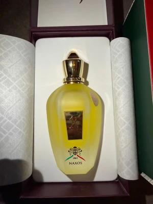 Xerjoff Naxos parfym 100ml - Lyxig parfym från Xerjoff, modell Naxos, i en elegant frostad gul flaska med gulddetaljer och exklusiv design. Flaskan har en guldfärgad kork och levereras i en snygg presentförpackning med vita och guldiga mönster. Perfekt för dig som gillar exklusiva dofter. KVITTO FINNS