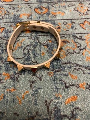 Valentino armband  - Snyggt vitt armband i läder med coola pyramidformade nitar i guld runt hela bandet. Justerbart spänne i guld för perfekt passform. Perfekt accessoar för dig som vill addera lite edge till din stil.