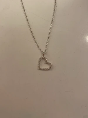 Silverhalsband med hjärta - Snyggt halsband i silverfärg med ett öppet hjärthänge dekorerat med små pärlor. Kedjan är tunn och har två inställningar. Endast använd fåtal gånger och aldrig duschat med. 