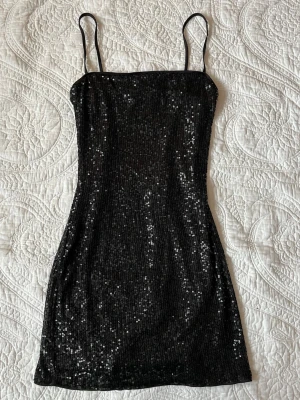 Svart paljettklänning med smala band - Snygg svart kortklänning med smala axelband och glittriga paljetter över hela klänningen. Figurnära passform och rak halsringning. Knappt använd så i mycket fint skick. Lappen är bortklippt då den lätt stack upp under användning.