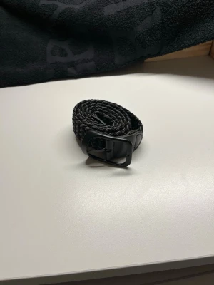 Svart flätat bälte med metallspänne - Snyggt svart flätat bälte med matchande svart metallspänne. Perfekt för att ge din outfit en clean och stilren touch. Bältet är flexibelt och passar till många olika looks.
