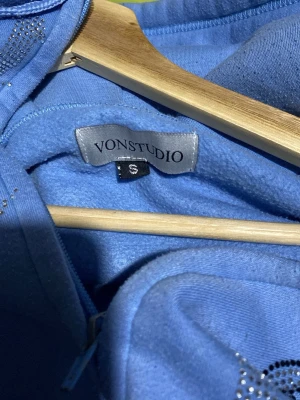 Blå hoodie med strass från Vonstudio - Blå zip-hoodie från Vonstudio med huva och fickor. Dekorerad med strassdetaljer och ett motiv av en ängel på framsidan. Tillverkad i mjukt material som känns skönt mot huden. Perfekt för dig som gillar streetwear och vill sticka ut.