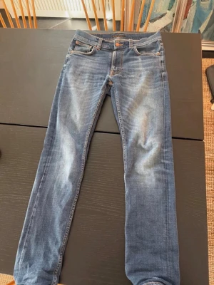 Blå slim fit jeans från Nudie Jeans - Säljer ett par blå slim fit jeans från Nudie Jeans med klassisk femficksmodell och snygga slitningar framtill. Jeansen har en mellanblå tvätt och detaljerade sömmar på bakfickorna. Tillverkade i bomull med en skön stretch för extra komfort.