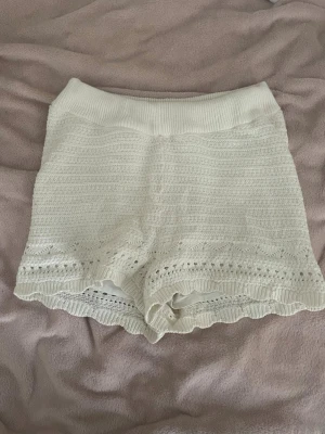 Vita stickade shorts med volangkant - Säljer ett par vita stickade shorts med bred resår i midjan och fina volangdetaljer längst ner. Shortsen har ett diskret hålmönster vid bensluten och känns både mjuka och luftiga. Perfekta för varma dagar eller som mysplagg hemma. Skriv för frågor!!