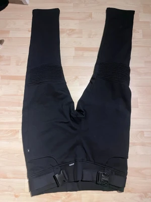 Svarta MC- byxor/ leggings med bälte - Säljer ett par svarta MC-inspirerade byxor med ribbade detaljer över knäna och justerbart bälte i midjan. Byxorna har raka ben och två bakfickor. Perfekta för dig som gillar en sportig och tuff stil. Storlek 6XL. Inbyggt skydd i. Nya endast testade men klippt av lappen. Katt finns i hemmet 