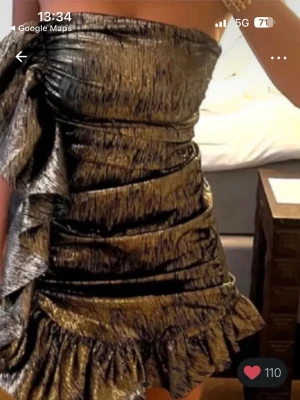 Guldig metallic kort klänning - Supersnygg kort klänning i guldig metallic med volangkant och stor rosett på sidan. Klänningen är axelbandslös och har en draperad look som ger en lyxig känsla. Perfekt för dig som vill sticka ut och glänsa.