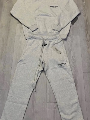Essentials tracksuit - Mysigt loungewear-set från Essentials i ljusgrått. Setet består av en hoodie med huva och tryckt logga på bröstet samt matchande mjukisbyxor med dragsko och logga. Perfekt för chill dagar hemma. Tillverkat i mjukt material för extra komfort.