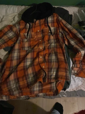 Orange-rutig overshirt med huva - Snygg overshirt i orange, svart och vitt rutmönster med svarta detaljer. Har en svart huva med dragsko, knäppning framtill och två bröstfickor. Perfekt för lager-på-lager och streetwear-stil.