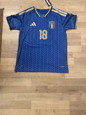 Italien fotbollströja Barella #18 Adidas Storlek S - Blå Italien fotbollströja från Adidas med guldiga detaljer och nummer 18, Barella på ryggen. Tröjan har korta ärmar, mönstrad design och Italiens landslagsmärke på bröstet. Perfekt för fotbollsfans som vill visa sitt stöd för landslaget. Storlek S