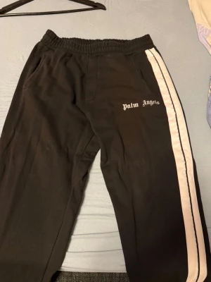 Palm Angels svarta mjukisbyxor - Svarta mjukisbyxor från Palm Angels med vit sidoremsa och logga tryckt på ena benet. Byxorna är tillverkade i 100% bomull. Perfekta för en avslappnad och trendig stil.