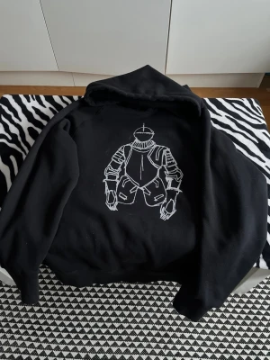 Svart hoodie från Number (N)ine - Svart hoodie från Number (N)ine med unik vit illustration av en riddarrustning på ryggen. Oversized passform och klassisk huva. Tillverkad av mjukt material som känns skönt mot huden. Perfekt för dig som gillar streetwear och statement-plagg.