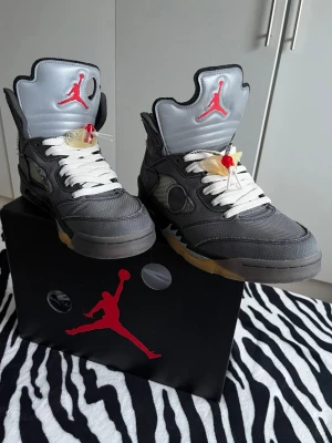 Air Jordan 5 Off-White  - Säljer ett par Air Jordan 5 Off-White sneakers i grått med vita skosnören, transparenta detaljer och röda Jumpman-loggor på plösen. Skorna har högt skaft och coola Off-White-texter på sidan. Sulan är delvis gul och har en unik design. Storlek 44