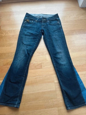 Blå bootcut jeans från G-Star - Säljer ett par snygga blå bootcut jeans från G-Star med klassisk femficksdesign och låg midja. Jeansen har slitningar och en cool tvätt som ger en avslappnad look. Tillverkade i denim med detaljerade sömmar och logga på bakfickan. Utsydda nertill i en ljusare jeans färg.