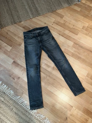 Grå Nudie jeans - Säljer ett par grå Nudie jeans i modellen Lean Dean med snygga slitningar och klassisk femficksmodell. Jeansen har en smal passform och är tillverkade i denim med stretch för extra komfort. Perfekta för en avslappnad och trendig look.