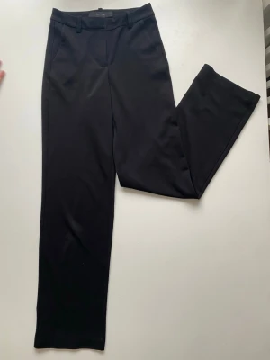 Svarta kostymbyxor från Vero Moda - Snygga svarta kostymbyxor från Vero Moda med raka ben och klassisk passform. Byxorna har hällor för skärp, två sidofickor och stängs med dragkedja och knapp. Perfekta till en stilren look och enkla att matcha med olika toppar.