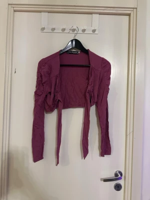 Lila bolero från Elvane - Trendig lila bolero från Elvane med långa ärmar och knytband framtill. Modellen är kort och har rynkade detaljer längs ärmarna för en cool och modern look. Perfekt att slänga över en topp för att lyfta outfiten.