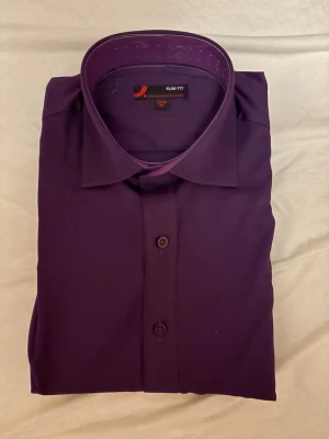 Lila slim fit skjorta från Dressmann - Snygg lila skjorta från Dressmann i slim fit-modell. Skjortan har klassisk krage, knappar framtill och är tillverkad i ett mjukt material som känns bekvämt mot huden. Perfekt för dig som vill ha en stilren look med lite färg.